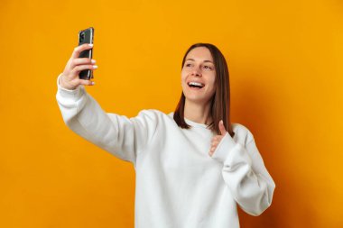 Joyful young woman is taking a selfie with her phone while showing thumb up over yellow background.