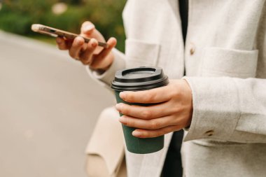 Close up shot of female hands holding phone and a cup of hot to go coffee outdoors.