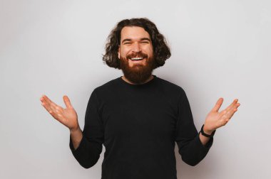 Young smiling bearded man wants to give you a hug and holds arms wide open. Studio shot over grey background.