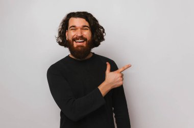 Studio portrait of a young happy bearded man is pointing aside at the light grey copy space.