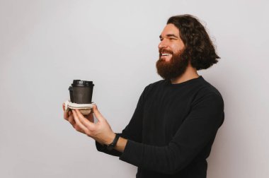 Handsome smiling bearded man is holding two take away cups of fresh coffee. Studio shot over grey background.