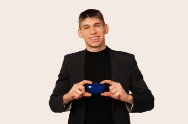 Handsome young man wearing black jacket recommends this blue credit card. Studio shot over white background.