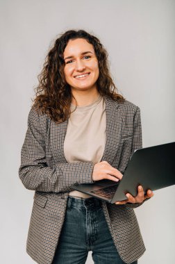 Vertical shot of a standing wide smiling woman holding an opened laptop. Studio photo over grey background.