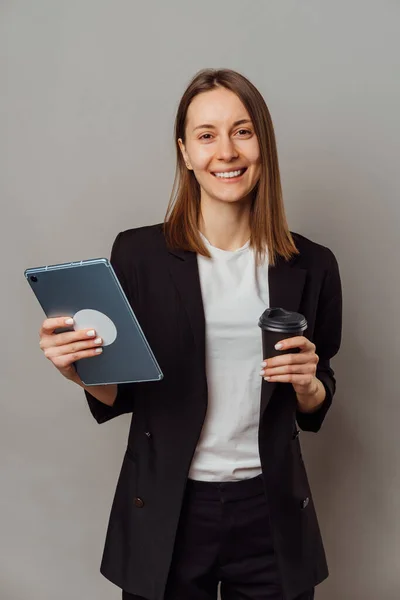 Confident woman wearing jacket holding a tablet and a to go cup of coffee over grey background.