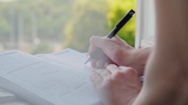Close up video of woman writing in her planner agenda in front of window.