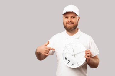 Cheerful delivery man wearing white points at a round clock he is holding. Studio shot over grey background.