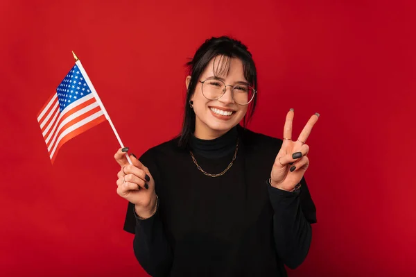 Wide smiling woman waves the USA flag for peace while showing the v gesture over red background.