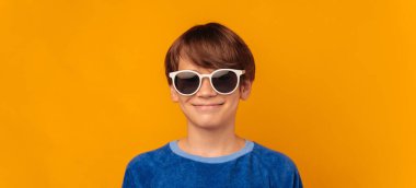 Banner shot of a shy smiling boy who is wearing cool white sunglasses over yellow background.