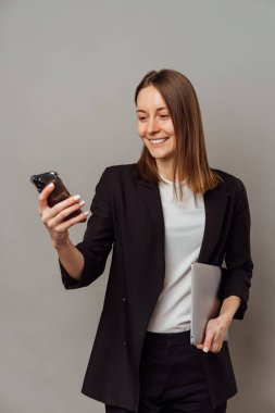Vertical studio shot of a smart smiling woman wearing jacket who is holding a laptop and her cellphone.
