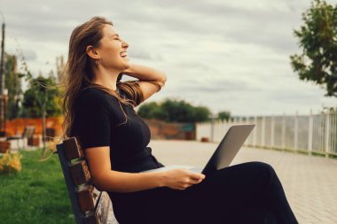 Young blonde woman is standing on a bench with laptop on legs and laughing outdoors.