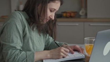 Video of young woman sitting at home in kitchen making some notes in agenda near.