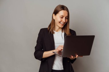 POrtrait of a cheerful smart woman is using laptop while holding one in a grey studio.