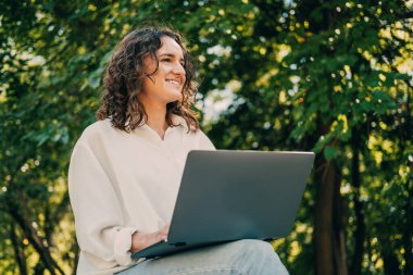 Young curly woman is sitting outdoors with an opened laptop on her knees in summer.