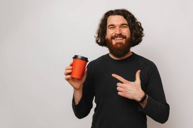 This take away coffee is awesome says a bearded man pointing at a red cup in a grey studio.