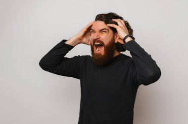 Angry bearded man is screaming and holding his head because of headache over grey background.