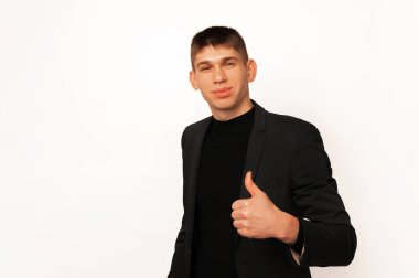 Handsome young smiling man wearing black jacket is holding a thumb up over white background.