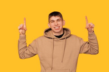 Cheerful young smiling man is pointing up above his head with both hands. Studio shot over yellow background.