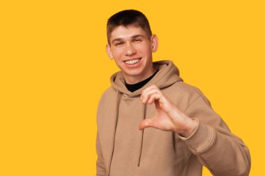 Wide smiling man is showing half a heart gesture over yellow background in a studio.