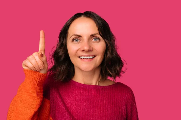 Look up says a mid age smiling brunette woman pointing above her head. Studio shot over pink background.