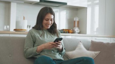 Footage of young cheerful woman sitting on sofa and typing in mobile phone at home.