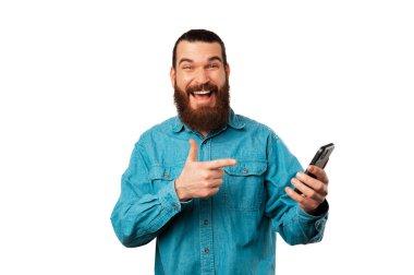 Yakışıklı, gülümseyen sakallı adam beyaz arka planda tuttuğu akıllı telefonu işaret ediyor..
