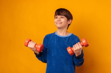 Yakışıklı, güçlü, gülümseyen genç çocuk sarı bir stüdyoda iki tane turuncu dambıl tutuyor..