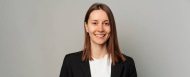 Gri arka planda, ceketli, gülümseyen bir kadının afiş boyunda portresi..