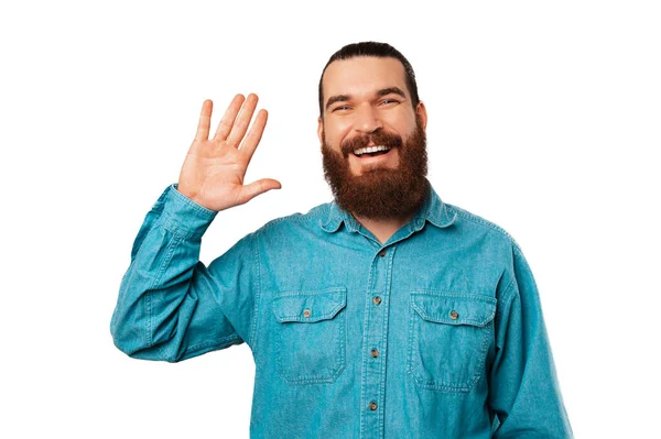 身穿蓝色衬衫的留着胡子的年轻人正在对着摄像机打招呼和挥手 白色背景的工作室肖像 — 图库照片
