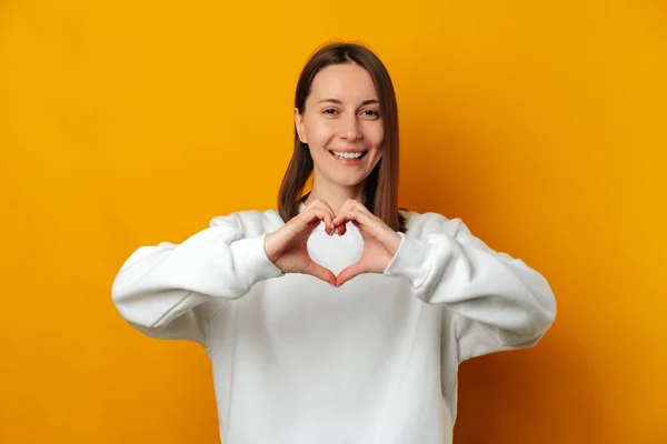 Dear women love yourselves says a girl showing a heart gesture over her chest. Studio shot over yellow background.