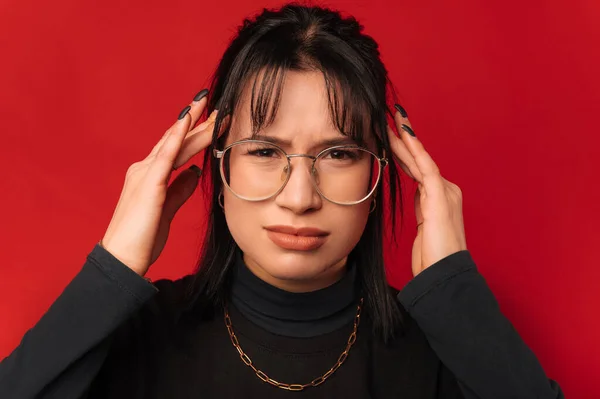 Young stylish frowning woman touching her head while having a bad headache over red background.