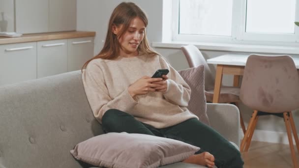 自宅のソファに座ってスマートフォンにメッセージを入力して笑う若い女性のビデオ — ストック動画