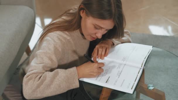 家に座って時間を計画しながら若い女性がプランナーの議題に何かを書いている映像 — ストック動画