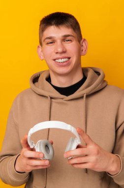 Sarı arka planda beyaz kulaklık tutan, kapüşonlu gülümseyen bir adamın dikey portresi..