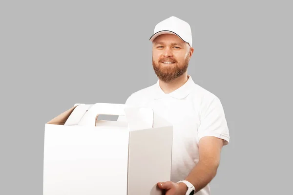 Smiling delivery man wearing white is holding a big cake delivery box over grey background.