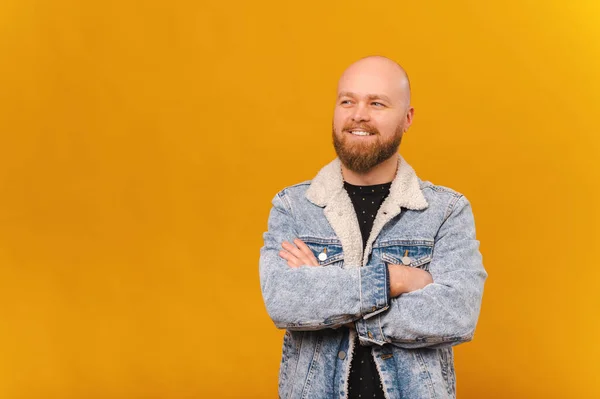 Confident smiling bearded bald man in jeans jacket is holding arms crossed over yellow background.