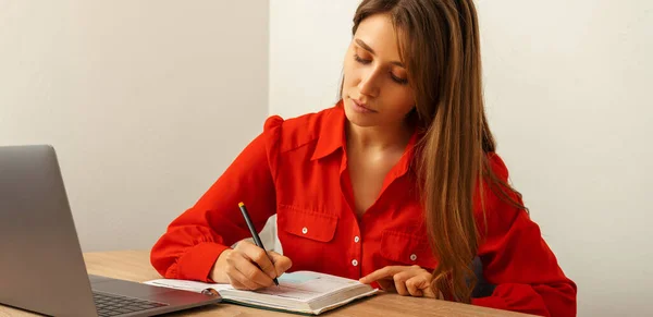 Banner size shot of a concentrated woman in red shirt writing in the journal at work.