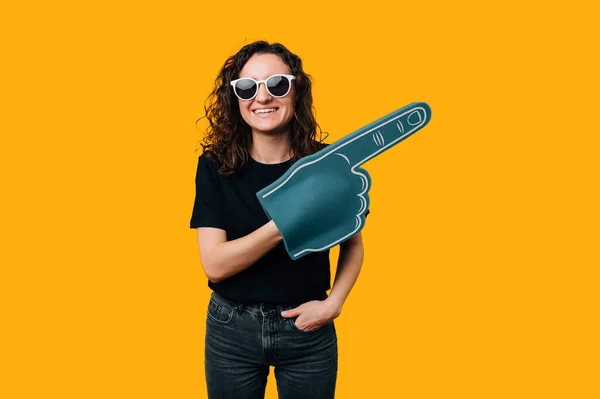 Excited young woman wearing glasses points aside while wearing fan foam glove over yellow background.