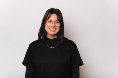 Gözlüklü neşeli siyah saçlı kadın gri arka planda kameraya gülümsüyor..