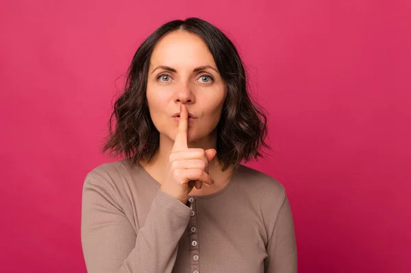 Beautiful brunette mid age woman is holding finger over her lips for silence over pink background.
