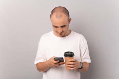  Kel adama bakmak ara verir internette sörf yapar ve gri arka planda kahvenin tadını çıkarır..