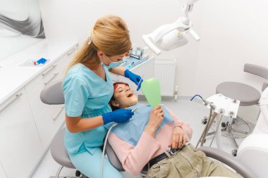 Genç kadın dişçi koltuğunda yatıyor ve küçük bir aynaya bakarken dişlerini yaptırıyor..