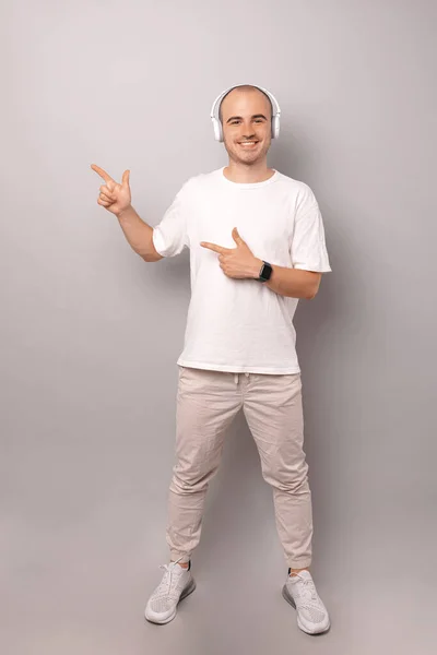 Full body shot of a bald man wearing headphones and pointing at copy space over grey background.