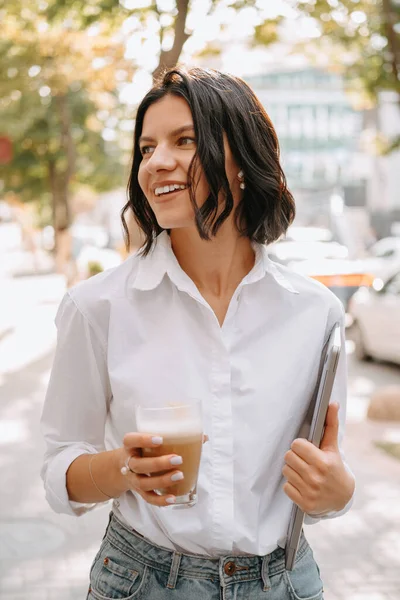 stock image Vertical shot of a woman walking outdoors while holding laptop and a glass of latte.