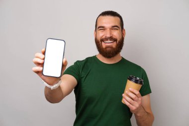 Heyecanlı genç sakallı adam, elinde kağıt bardak tutarken gri arkaplanda telefon ekranı gösteriyor..