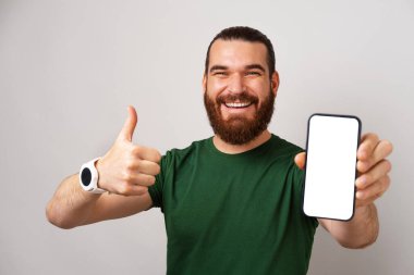 Yakışıklı sakallı adam elinde boş bir telefon ekranı tutuyor ve gri arka planda baş parmağını gösteriyor..