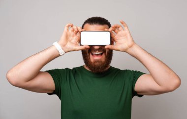 Mest sakallı adam gözlerini telefonuyla kapatıyor. Gri arkaplan üzerinde boş bir ekran var..