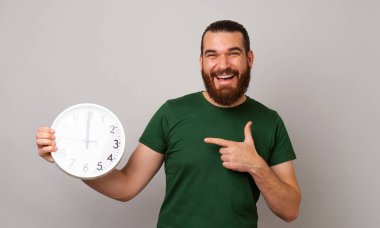 Yeşil tişört giyen genç sakallı adam gri arka planda dururken yuvarlak beyaz bir saati işaret ediyor..