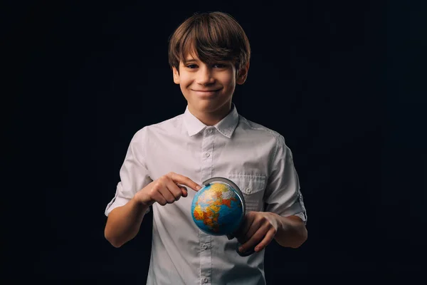 Young teen boy wearing white shirt is holding and pointing on a small Earth globe.