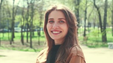 Şehir parkında dikilirken kameraya bakıp gülümseyen neşeli genç bir kadının portresi..