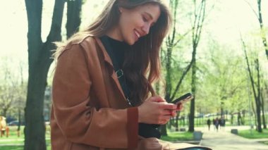 Şehir parkında bankta oturan ve baharda akıllı telefon kullanan genç bir kadının videosu..